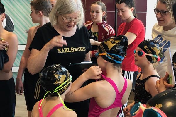 Eine Trainerin erklärt Schwimmanfängern mit Badekappen und Schwimmbrillen etwas am Beckenrand.