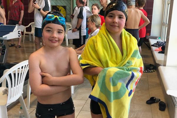 Zwei Kinder in Badesachen stehen in einer Schwimmhalle, eines mit Schwimmbrille, das andere in ein Handtuch gehüllt.