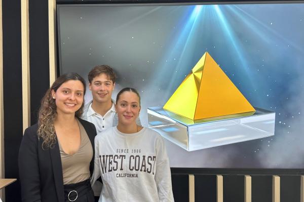Drei Personen stehen vor einem Bildschirm mit einer gelben Pyramide auf einer transparenten Basis.