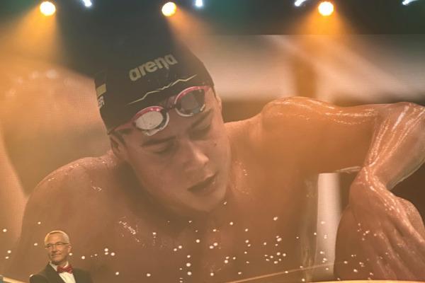Mann mit Schwimmbrille auf der Bühne, im Hintergrund ein Bildschirm mit einem schwimmenden Athleten.