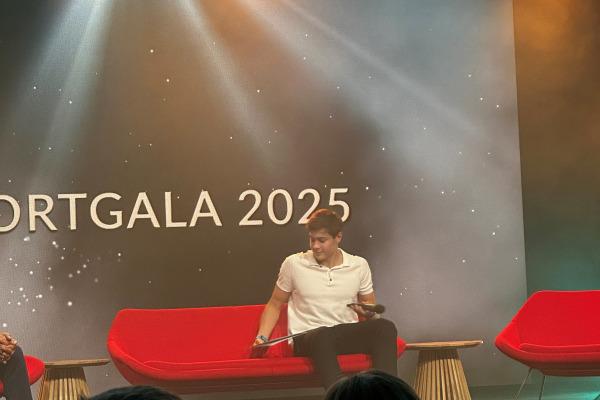 Mann in weißem Shirt sitzt auf rotem Sofa bei der Sportgala 2025, mit Lichtern und einer Präsentationsleinwand im Hintergrund.