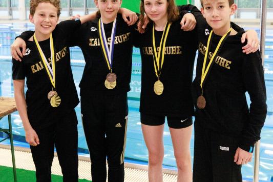 Vier junge Schwimmer stehen lächelnd zusammen, tragen Medaillen und schwarze Trikots mit der Aufschrift „Karlsruhe“.