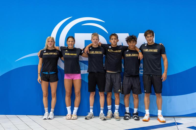 Sechs Schwimmer in schwarzen T-Shirts mit "Karlsruhe" stehen lächelnd vor blauer Wand in einem Schwimmbecken.