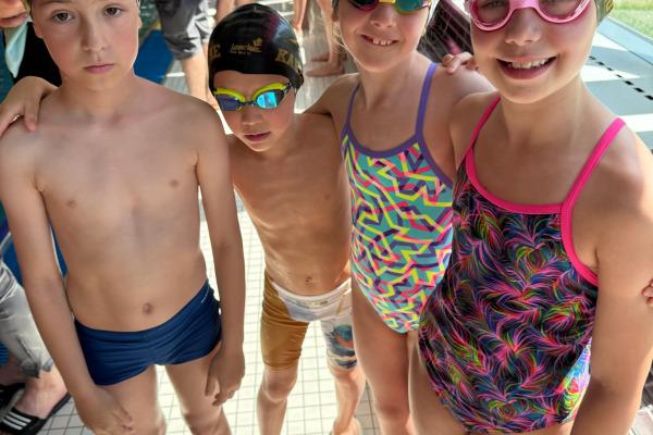 Vier Kinder in Badebekleidung und Schwimmkappen stehen zusammen am Poolrand, fröhlich lächelnd für ein Foto.