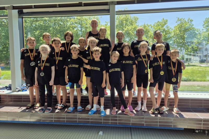 Gruppe von Kindern in Sportkleidung mit Medaillen, posierend vor einem Fenster in einem Schwimmbad.