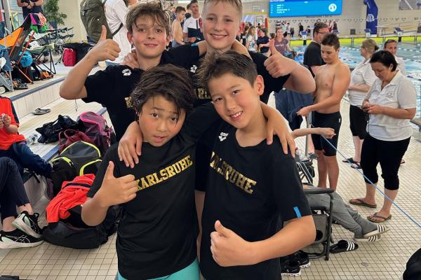 Vier Kinder in schwarz-gelben T-Shirts mit "Karlsruhe" posieren fröhlich am Beckenrand eines Schwimmbads.