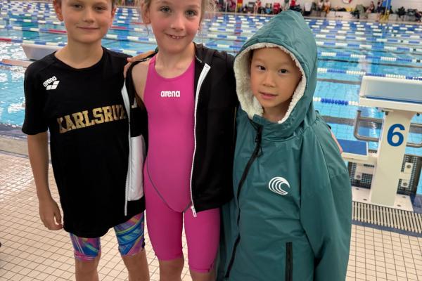 Drei Kinder stehen am Schwimmbecken: ein Junge in schwarzem Shirt, ein Mädchen in pinkem Badeanzug und ein Junge in grünem Poncho.