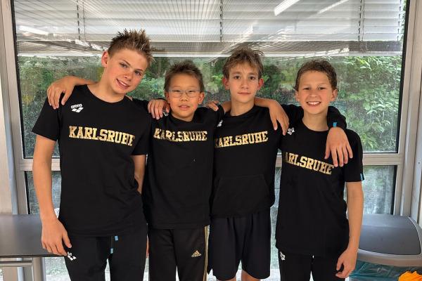 Vier Kinder stehen lächelnd zusammen in schwarzen T-Shirts mit der Aufschrift "Karlsruhe" vor einem Fenster.