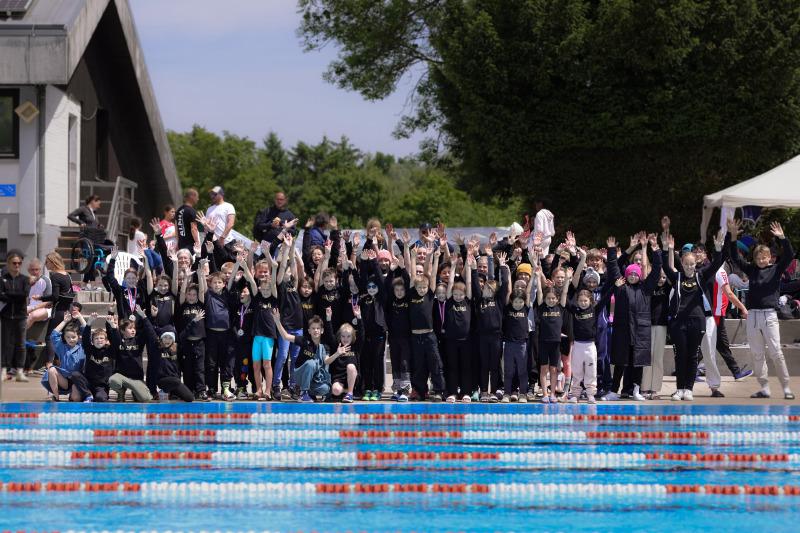 Gruppenselfie von Kindern und Erwachsenen am Schwimmbad, alle mit erhobenen Händen und Medaillen um den Hals.