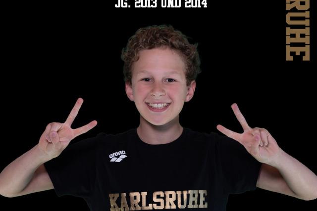 Junge mit lockigem Haar macht Peace-Zeichen, trägt schwarzes T-Shirt mit "Karlsruhe" und Hintergrund in Schwarz.
