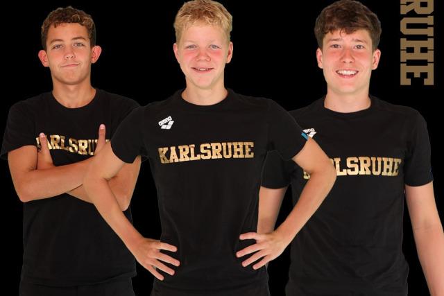Drei junge Sportler in schwarzen T-Shirts mit "Karlsruhe" posieren für ein Gruppenbild. Hintergrund: Stadtansicht Rostock.