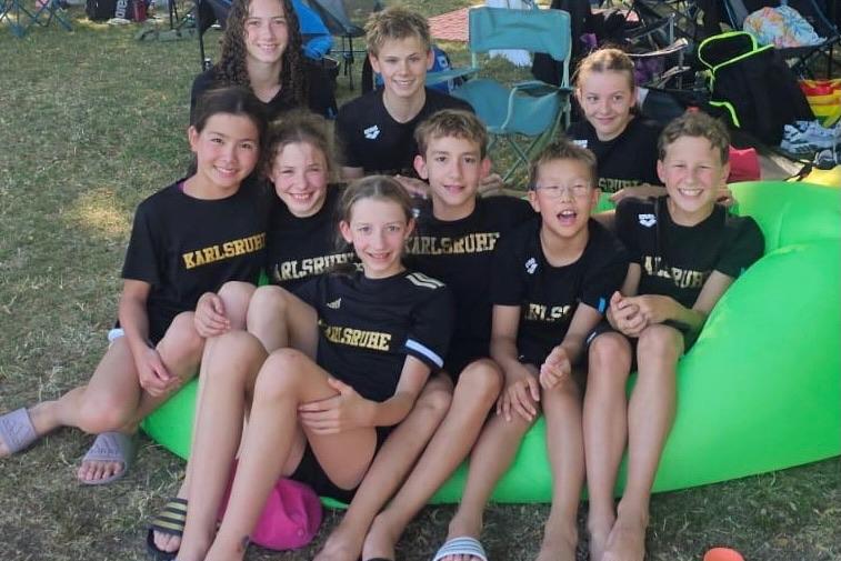 Gruppe von neun Kindern in T-Shirts mit Aufdruck „Karlsruhe“ auf einer Wiese, umgeben von Campingstühlen.