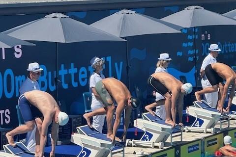Wettkampfschwimmer in Startpositionen an Beckenrand vor einer Schwimmübertragung mit mehreren Lane-Markierungen.