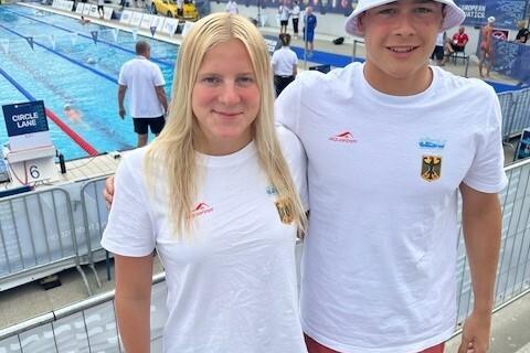 Zwei jugendliche Schwimmer posieren am Beckenrand eines Schwimmwettbewerbs, beide tragen weiße T-Shirts und tragen sportliche Kleidung.