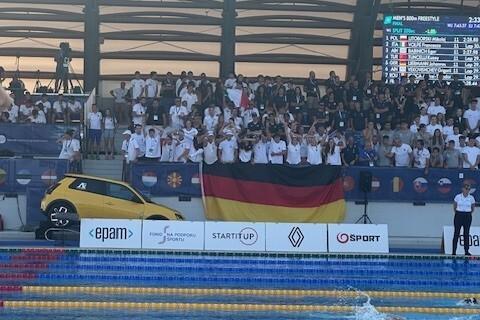 Zuschauer mit deutscher Fahne sitzen an einem Schwimmbecken während eines Wettkampfs, im Hintergrund ist eine Anzeigetafel sichtbar.