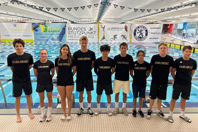 Gruppe junger Schwimmer in schwarzen T-Shirts mit „Karlsruhe“-Aufdruck am Beckenrand eines Schwimmbads.