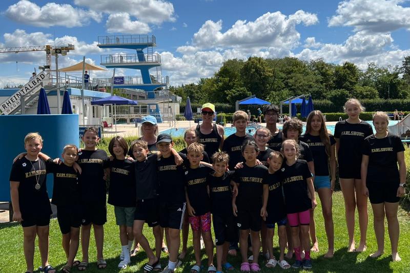 Gruppe von Kindern und Betreuern in einheitlichen T-Shirts vor einem Schwimmbad mit Sprungturm und blauen Liegen.