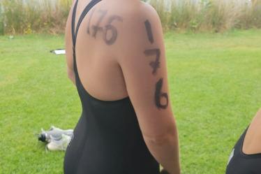 Wettkampfschwimmerin in schwarzem Anzug mit Schwimmhaube, Zahlen auf dem Arm, steht am Ufer eines Gewässers.