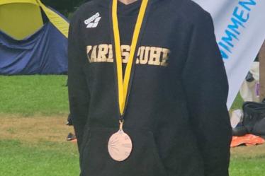 Mädchen in schwarzem Hoodie mit Medaille steht vor Zelt und Banner im Freien.