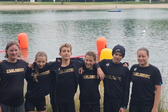 Sechs Kinder in Schwarz mit “Karlsruhe”-Aufdruck stehen am Ufer eines Gewässers, hinter ihnen orange Markierungen im Wasser.