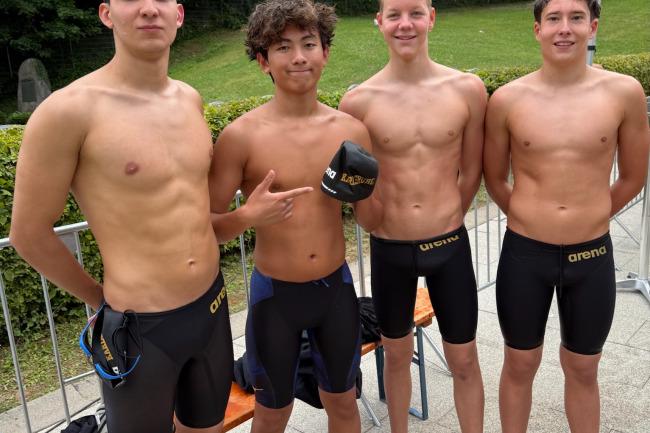 Vier junge Schwimmer stehen auf einer Freiluftanlage, tragen Badeanzüge und Halbmützen, im Hintergrund Bäume.