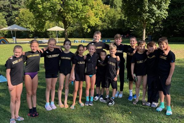 Gruppe von Kindern in schwarzen T-Shirts mit „Karlsruhe“ auf einer Wiese, umgeben von Bäumen und Sonnenschirmen.
