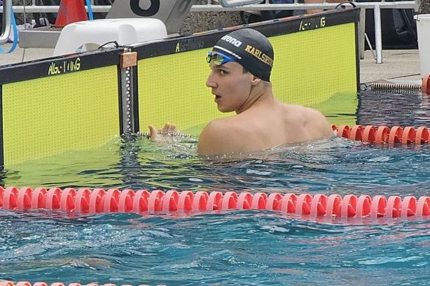 Schwimmer in der Badehose mit Kappe schaut über den Beckenrand, Schwimmhalle mit gepunkteten Schwimmbahnen im Hintergrund.
