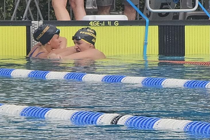 Zwei Schwimmerinnen in Badekappen umarmen sich im Wasser eines Schwimmbeckens, im Hintergrund stehen Zuschauer.