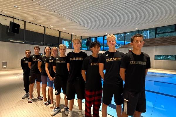 Gruppierung von zehn jungen Sportlern in schwarzen T-Shirts mit "Karlsruhe" am Beckenrand eines Schwimmbads.