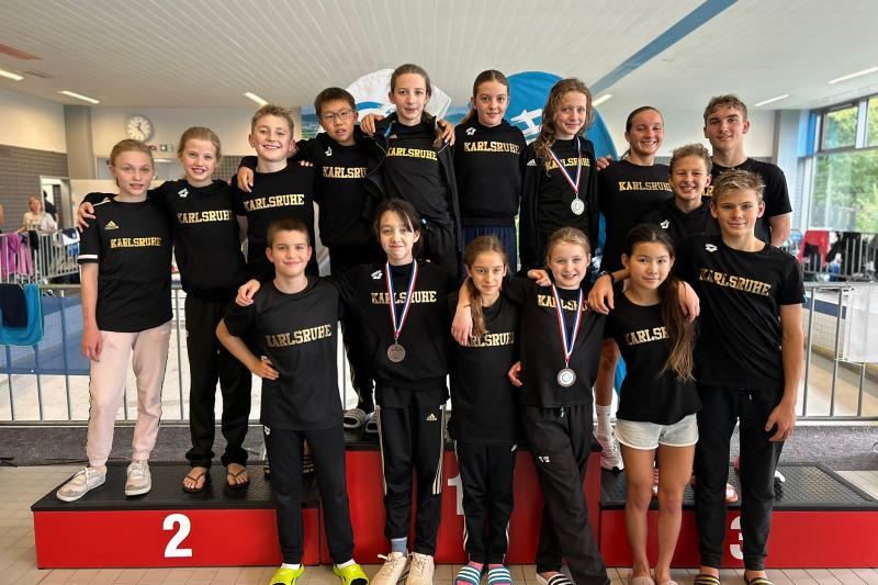 Gruppe von Schwimmsportlern in schwarzen T-Shirts mit der Aufschrift "Karlsruhe" auf dem Siegerpodest nach einem Wettkampf.