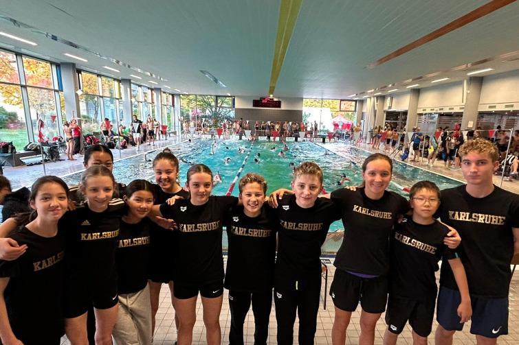 Gruppenshot von Schwimmern in einheitlichen schwarzen Shirts mit "Karlsruhe" vor einem Schwimmbecken mit Wettkämpfen.