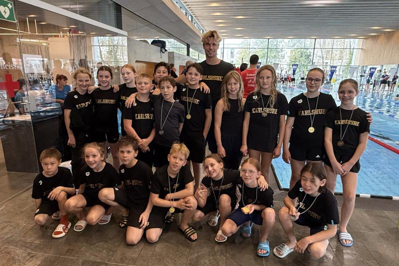 Gruppe von Schwimmern in schwarzen T-Shirts mit Aufschrift „Karlsruhe“ und Medaillen in einem Schwimmbad.