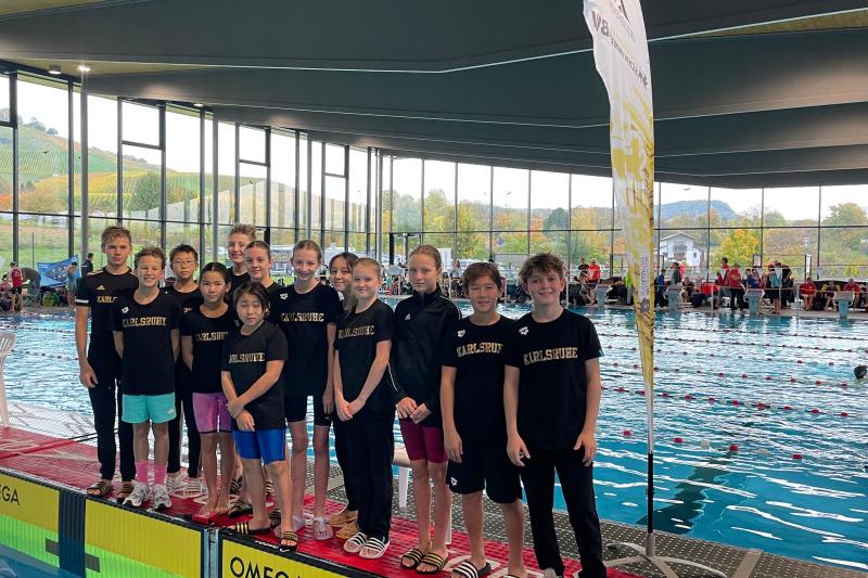 Gruppe junger Schwimmer in einheitlichen T-Shirts mit „Karlsruhe“ stehen am Beckenrand in einem modernen Schwimmbad.