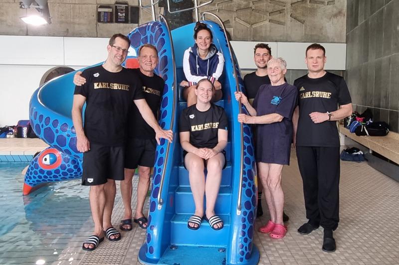 Gruppe von sechs Personen vor einer blauen Wasserrutsche in einem Hallenbad, lächelnd und in Sportkleidung.