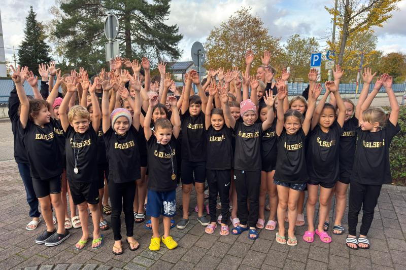 Gruppierung von Kindern in schwarzen T-Shirts mit erhobenen Händen, umgeben von Bäumen und einer Parkplatzschild.