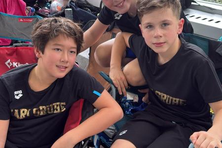 Drei Jungen in Sportkleidung sitzen lächelnd auf Campingstühlen in einer Sporteinrichtung.