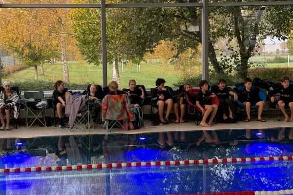 Gruppe von Schwimmern sitzt auf Stühlen am Beckenrand eines Pools, umgeben von herbstlicher Natur.
