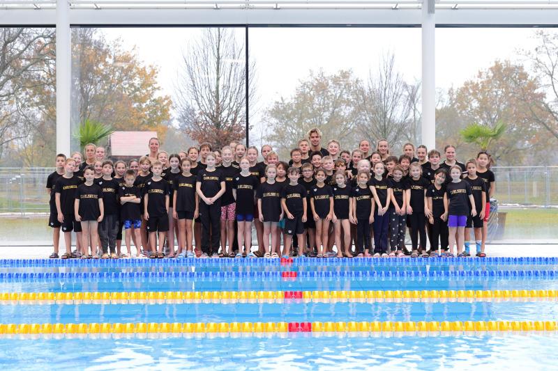 Gruppenfoto von Schwimmern in schwarzen T-Shirts am Beckenrand eines Hallenbads, umgeben von bunten Wasseranlagen.