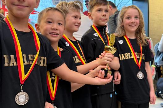 Gruppe von fünf Kindern in Sportkleidung mit Medaillen und einem Pokal in der Hand, lächelnd vor einem Hintergrund.