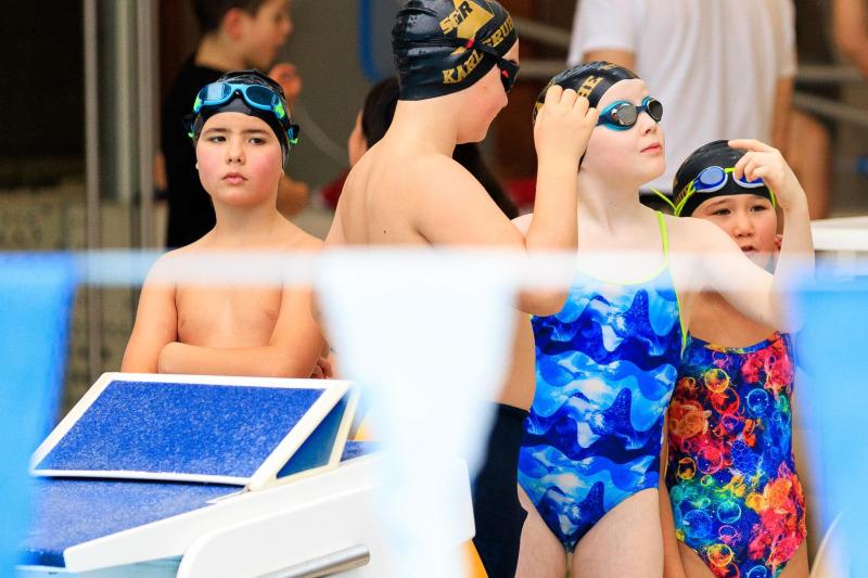 Vier Kinder in Badekleidung und Schwimmbrillen stehen am Beckenrand, bereit für den Wettkampf im Schwimmbad.