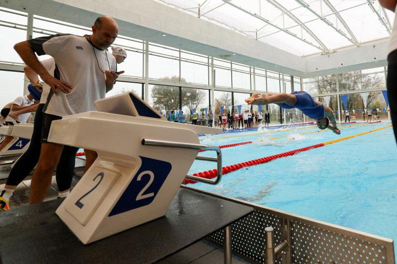 Schwimmer springt von Startblock 2 ins Wasser, während Trainer und Zuschauer am Beckenrand beobachten.
