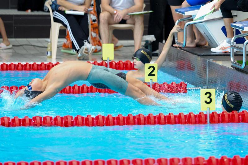 Junger Schwimmer in Hautfarbe mit blauen Badekappe springt ins Wasser, während andere Wettkämpfer warten.