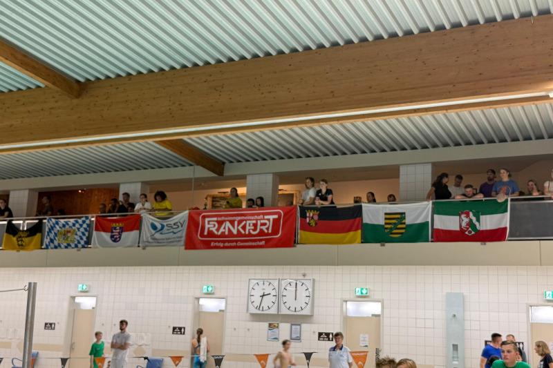 Blick auf Zuschauer mit Fahnen an einer Galerie über einem Schwimmbecken, mit Sportlern im Vordergrund.