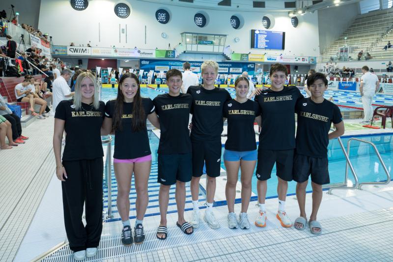 Gruppe von Schwimmern in einheitlichen schwarzen Shirts mit dem Aufdruck "Karlsruhe" vor einem Wettkampfbecken.
