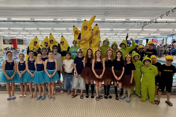 Gruppennfoto von Kindern in lustigen Kostümen, darunter Bananen und Ungeheuer, vor einem Schwimmbecken.