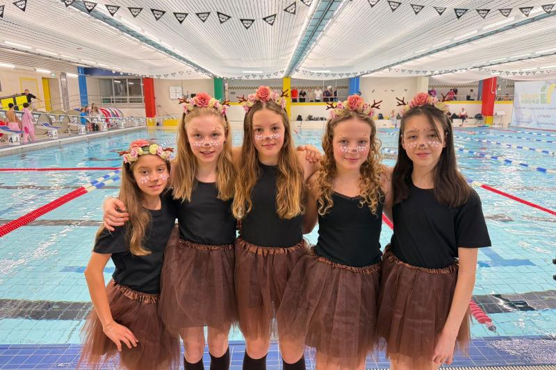 Fünf Mädchen in schwarzen T-Shirts und braunen Tutus posieren am Beckenrand eines Schwimmbads mit Blumenkränzen im Haar.