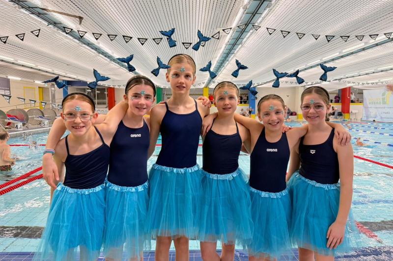 Sechs Mädchen im Schwimmanzug mit blauen Tüllröcken und Glitzer schminken sich am Schwimmbeckenrand.
