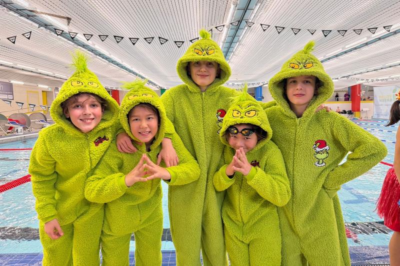 Fünf Kinder in grünen Grinch-Mänteln posieren lächelnd am Beckenrand eines Schwimmbades.