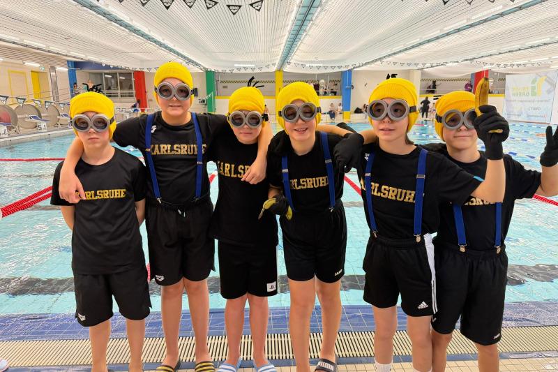 Sechs Kinder in Schwimmbekleidung mit gelben Mützen und Schwimmbrillen posieren am Beckenrand eines Schwimmbads.