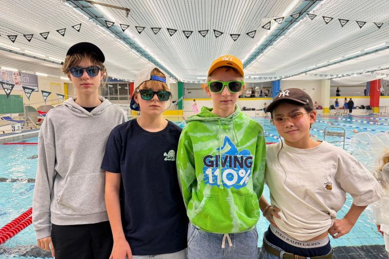 Vier Jugendliche stehen am Beckenrand eines Schwimmbads, tragen Sonnenbrillen und lässige Kleidung.